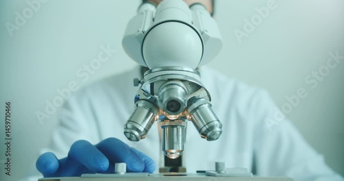 Close-up of biologist gloved hands using microscope in sterile laboratory. Vibrant blue gloves highlight precise scientific work against monochromatic lab backdrop