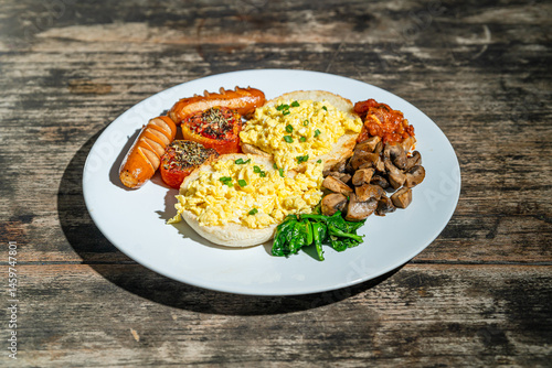 Big breakfast with bread toast, scrambled egg, sausages, roasted tomato, baked bean, sauted mushroom, and wilted spinach on white plate. Isolated by wooden table. Food Photography