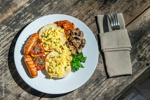 Big breakfast with bread toast, scrambled egg, sausages, roasted tomato, baked bean, sauted mushroom, and wilted spinach on white plate. Isolated by wooden table. Food Photography