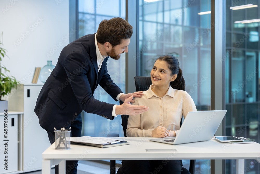 A businesswoman sits at her desk, listening attentively while her colleague offers guidance, inside a bright office setting.