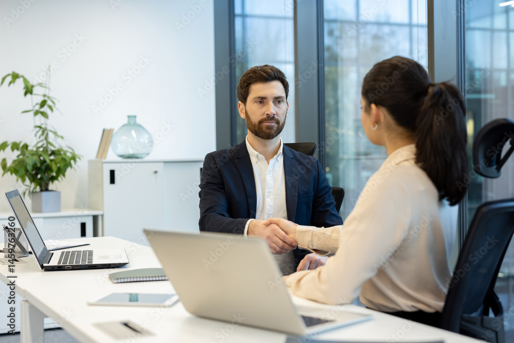 A business handshake seals a deal between two individuals seated at a modern office desk with laptops. The scene shows professional agreement and teamwork.