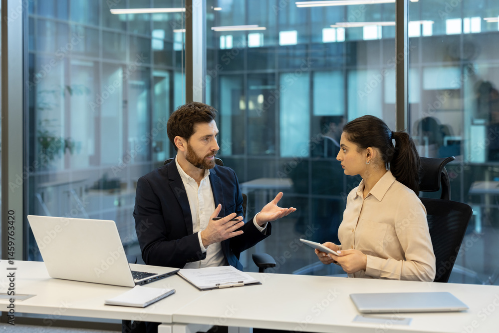 A businessman and businesswoman collaborate in an office setting, discussing using a laptop, tablet, and documents. The environment suggests a modern and professional workspace.