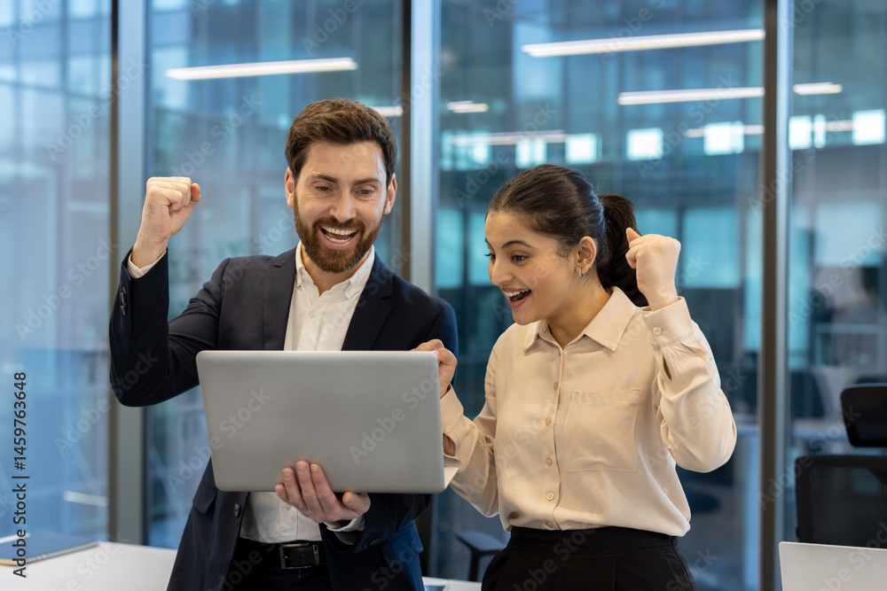A man and woman celebrate success while looking at a laptop in an office setting, happy and excited. The building's modern interior is visible.