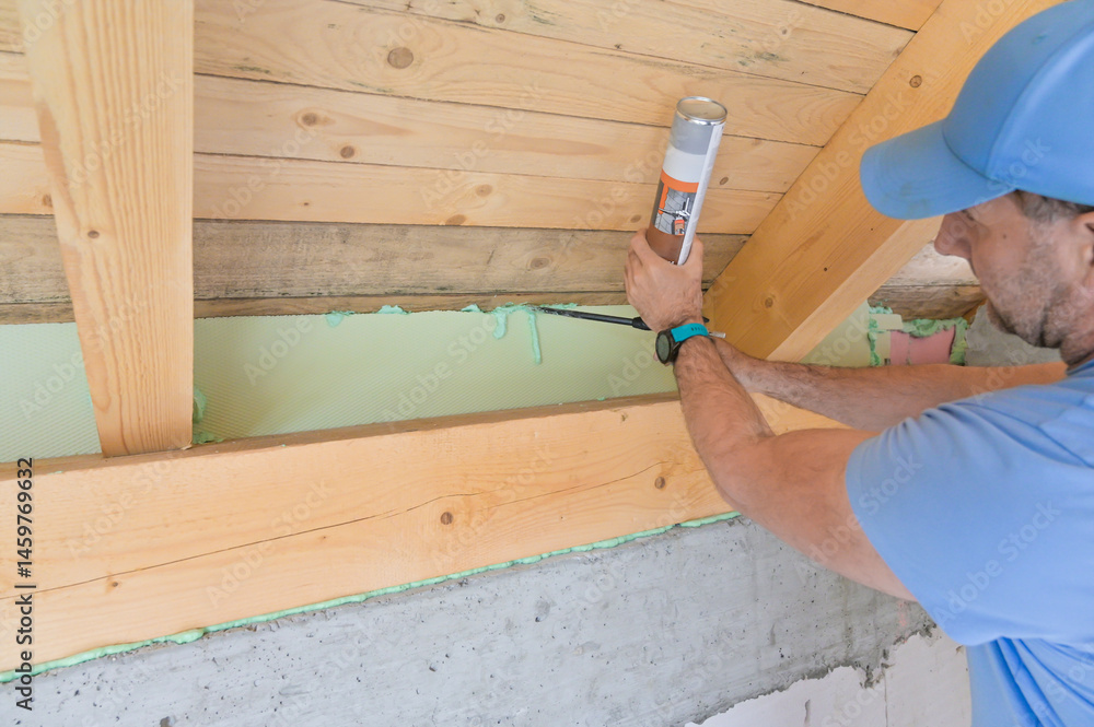 A man uses a caulking gun to apply insulation along a wooden beam in a home renovation project, highlighting the importance of energy efficiency and construction skills