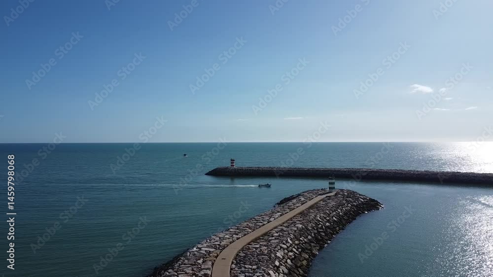 A small boat navigates the calm waters of Quarteira, Portugal, passing between the twin lighthouses that guard the harbour. A small boat navigates the calm waters of Quarteira, Portugal, passing between the twin lighthouses that guard the harbour.