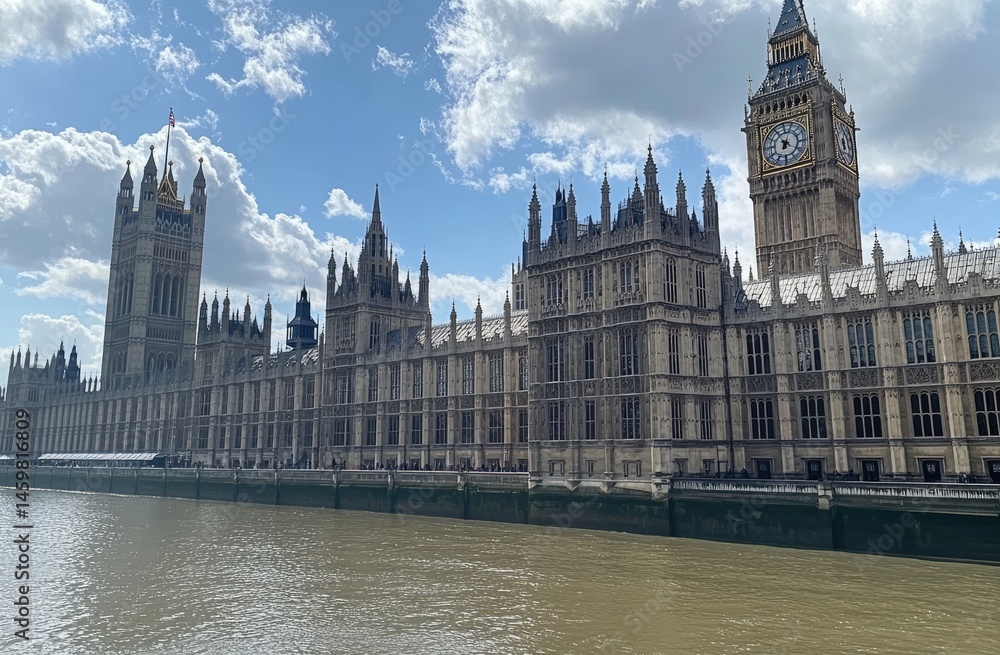 Fototapeta premium Historic British parliament building alongside a river, under a partly cloudy sky. Tall towers and intricate architectural details are visible