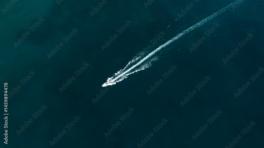 Aerial zenithal view of a motorboat crossing the blue sea. Its passage leaves a white trail on the surface of the water.