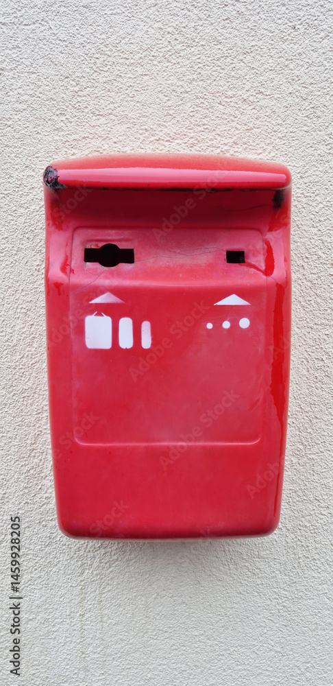 A bright red, slightly weathered metal box with white symbols is mounted on a textured light-colored wall, indicating a collection point for old batteries