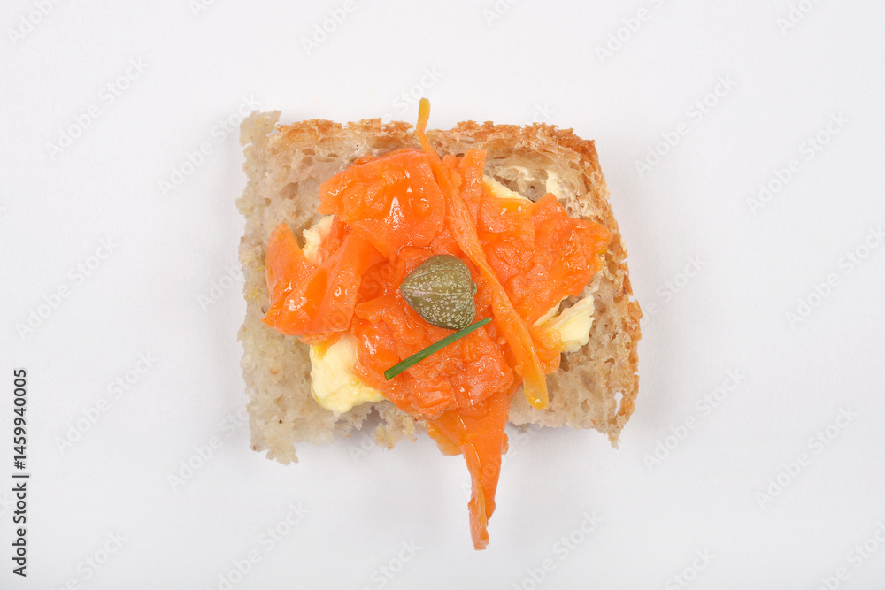 A piece of bread topped with vegan carrot lox, cream cheese, and a caper is centered on a white background