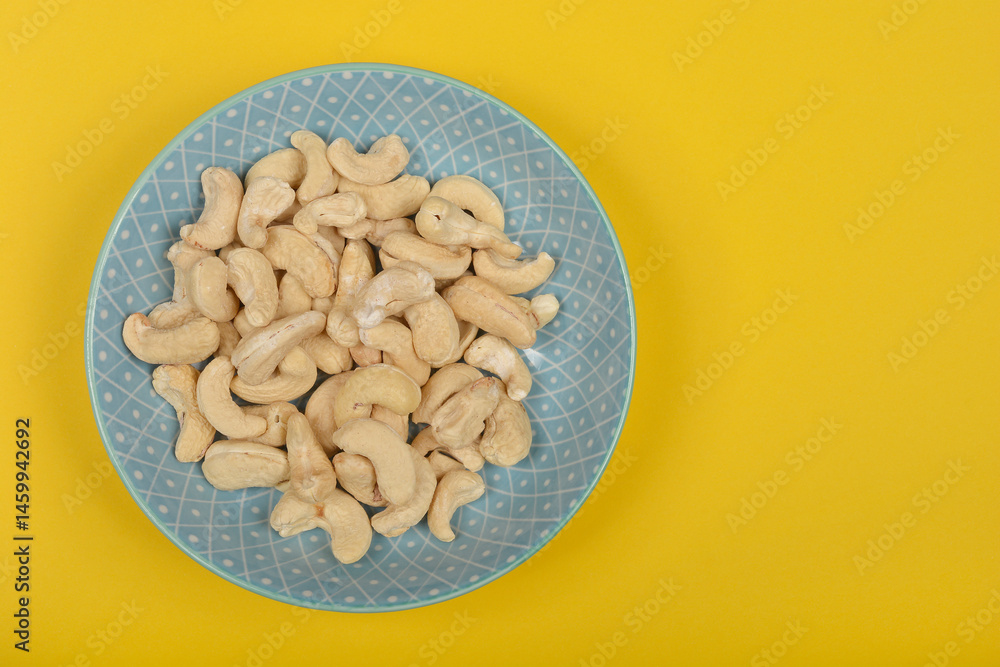 A light blue oval plate filled with raw cashew nuts is positioned on a bright yellow background