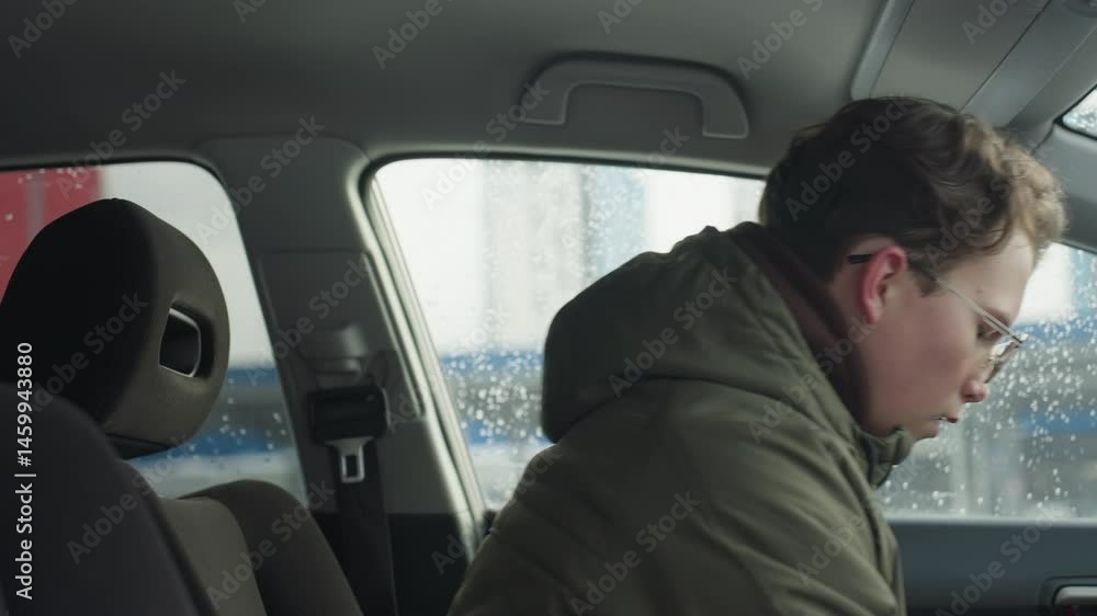 close up side view of young man in winter jacket adjusting car seat forward in preparation to drive with raindrops on window and blurred building visible outside during cold season close up side view of young man in winter jacket adjusting car seat forward in preparation to drive with raindrops on window and blurred building visible outside during cold season