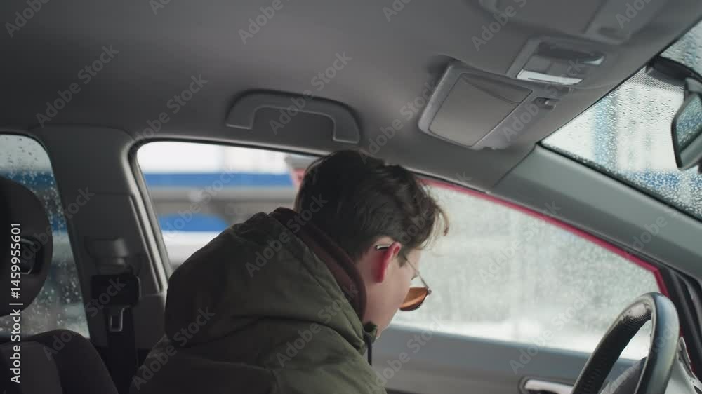 Young boy in winter jacket exits car during cold season with water droplets on window, urban background softly blurred through glass, interior details visible including seat and roof handle Young boy in winter jacket exits car during cold season with water droplets on window, urban background softly blurred through glass, interior details visible including seat and roof handle