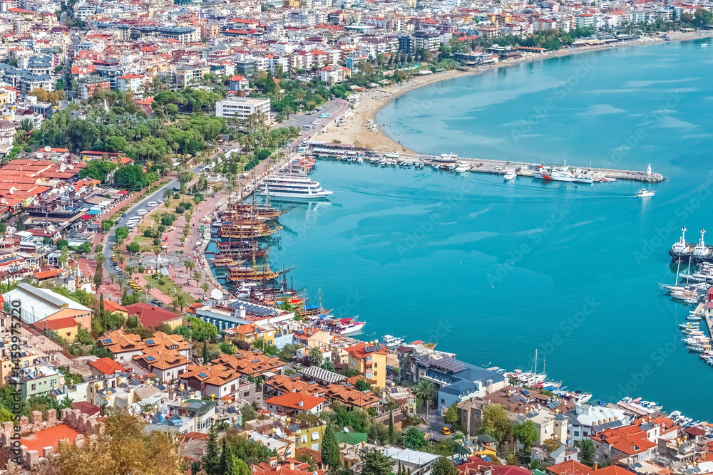 Fototapeta premium Aerial view of Alanya's harbor with colorful pirate ships docked along the promenade and nearby beach. The bustling coastal area offers a beautiful panorama of the city and the Mediterranean Sea