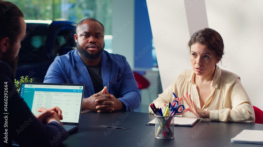 Customers in showroom completing credit application after being sold car insurance by salesman. Clients in dealership signing financing paperwork after buying extra vehicle warranty, camera A