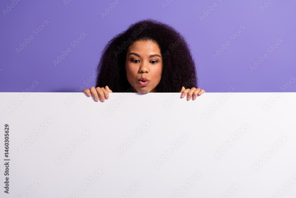 Curious young woman peeking over a white blank board with a playful expression against a purple backdrop