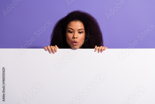 Obraz na płótnie Curious young woman peeking over a white blank board with a playful expression against a purple backdrop w salonie