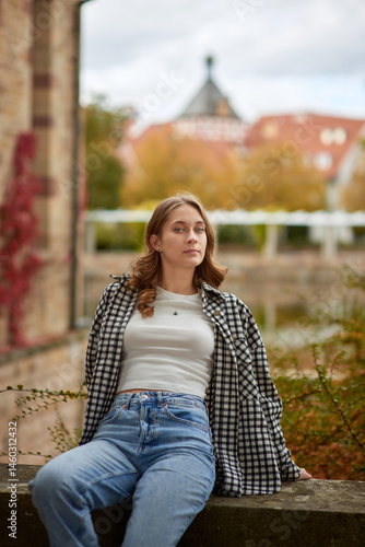 Young woman in casual autumn outfit relaxing in a historic European city park with medieval architecture in the background.