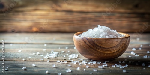Wooden bowl filled with coarse sea salt crystals scattered on rustic wooden surface