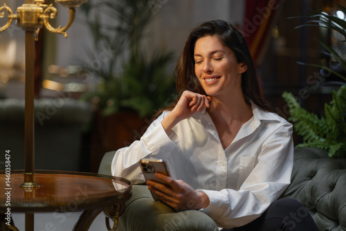 Smiling young businesswoman using smartphone sitting on cushioned chair in hotel lobby. Woman in white shirt and appears relaxed. Warm lighting create cozy atmosphere