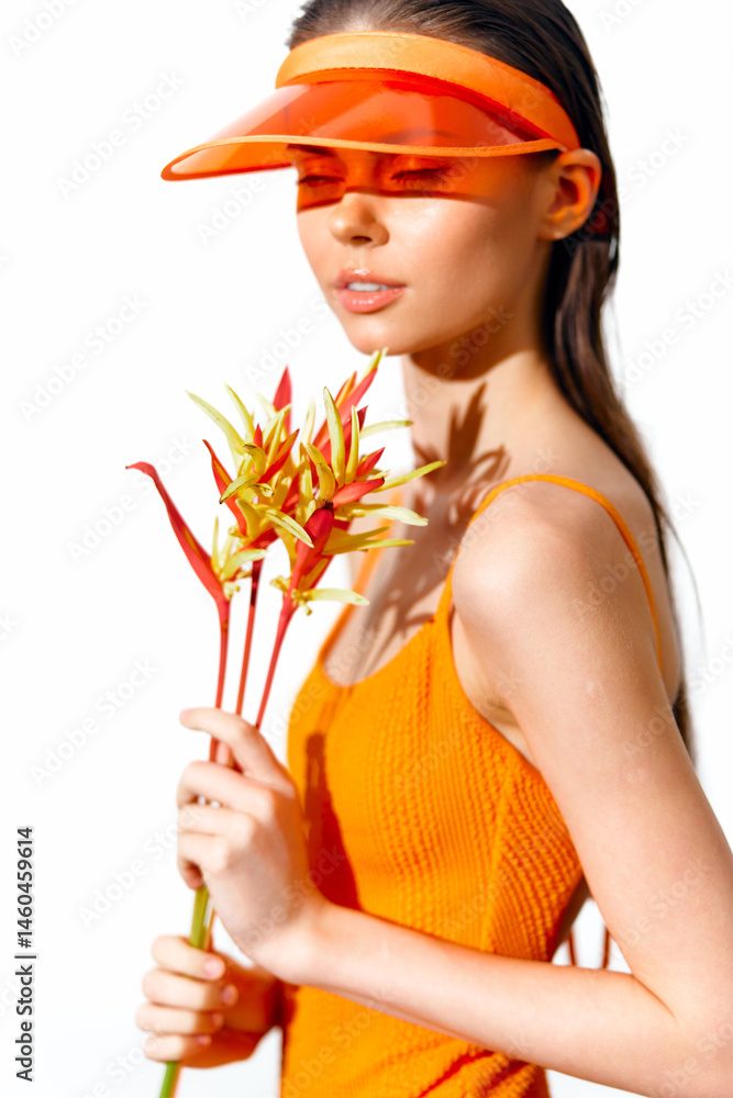 Young woman in bright orange dress and visor holding tropical flowers, exuding joyfulness and summer vibes.