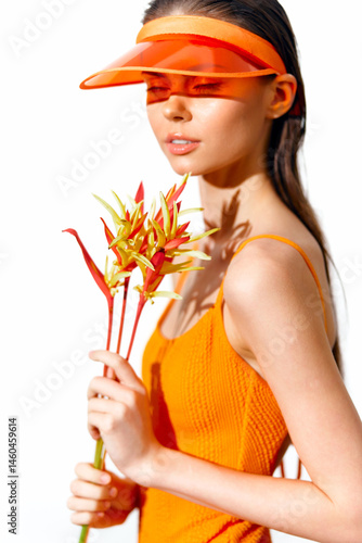 Obraz na płótnie Young woman in bright orange dress and visor holding tropical flowers, exuding joyfulness and summer vibes. w salonie