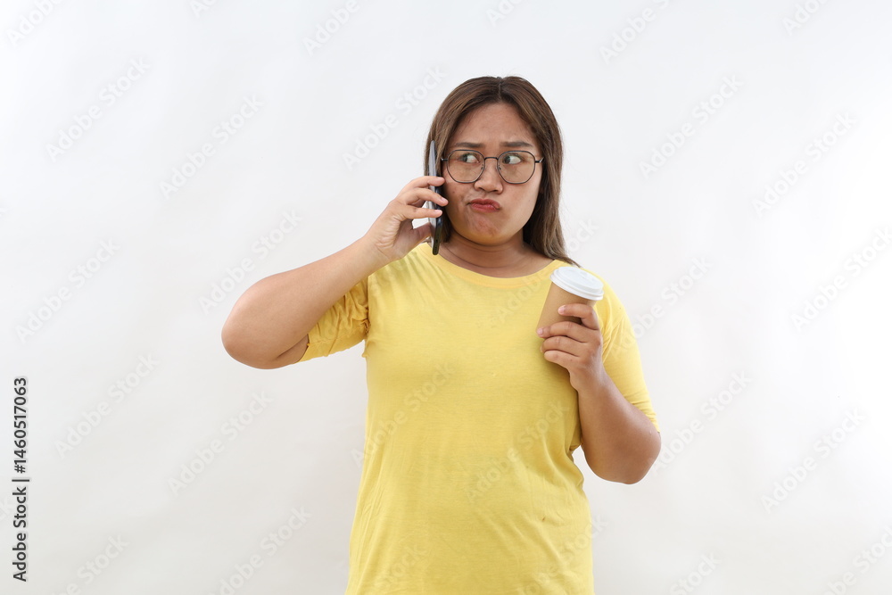 Asian female makes phone call, tired of waiting someone answers, keeps cellular near ear, drinks takeaway coffee, dressed in casual t-shirt, poses indoor. Boring conversation Asian female makes phone call, tired of waiting someone answers, keeps cellular near ear, drinks takeaway coffee, dressed in casual t-shirt, poses indoor. Boring conversation
