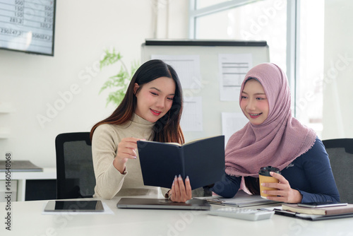 Two Young Asian Women Collaborate on Muslim Business Strategy Reading Book Holding Coffee Cup in Modern Office Positive Meeting Teamwork