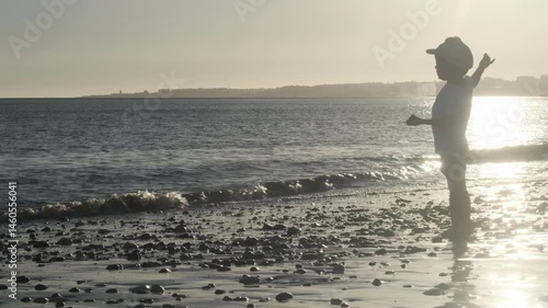 Backlit silhouette of young child playing along sandy shoreline during golden hour sunset, experiencing carefree childhood moment near ocean waters in portugal