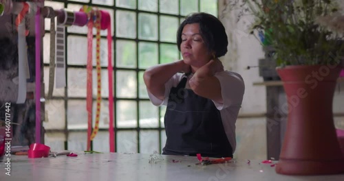 Female florist stretching neck and relaxing after work at rustic flower shop, small business owner taking a break to recover from busy workday