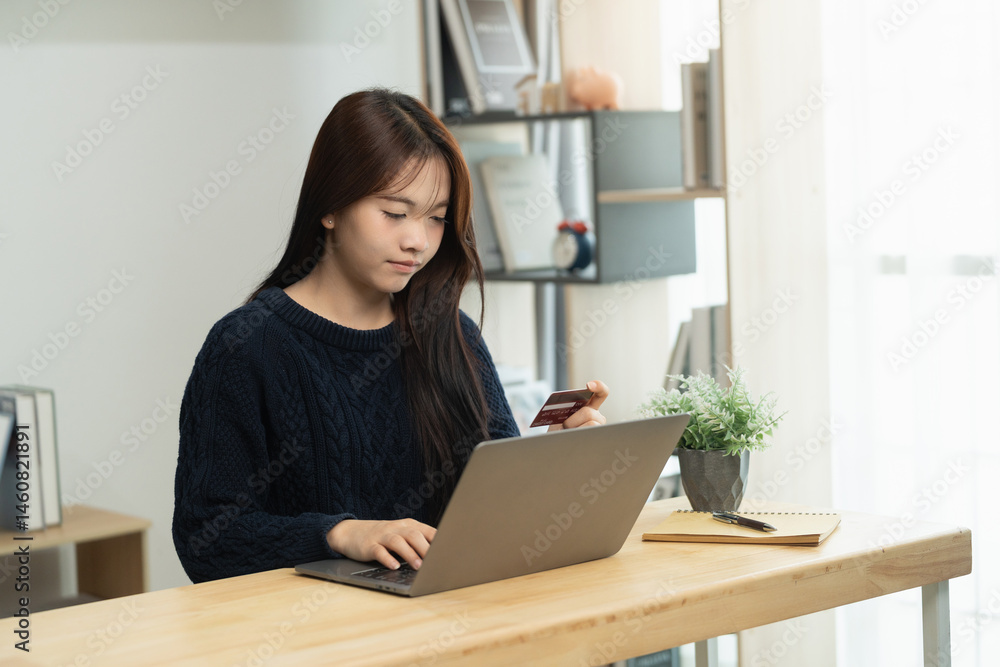 Young Asian woman sits at desk using laptop for online shopping, holding credit card, casual home office setting, soft natural light, focused expression, technology lifestyle Young Asian woman sits at desk using laptop for online shopping, holding credit card, casual home office setting, soft natural light, focused expression, technology lifestyle