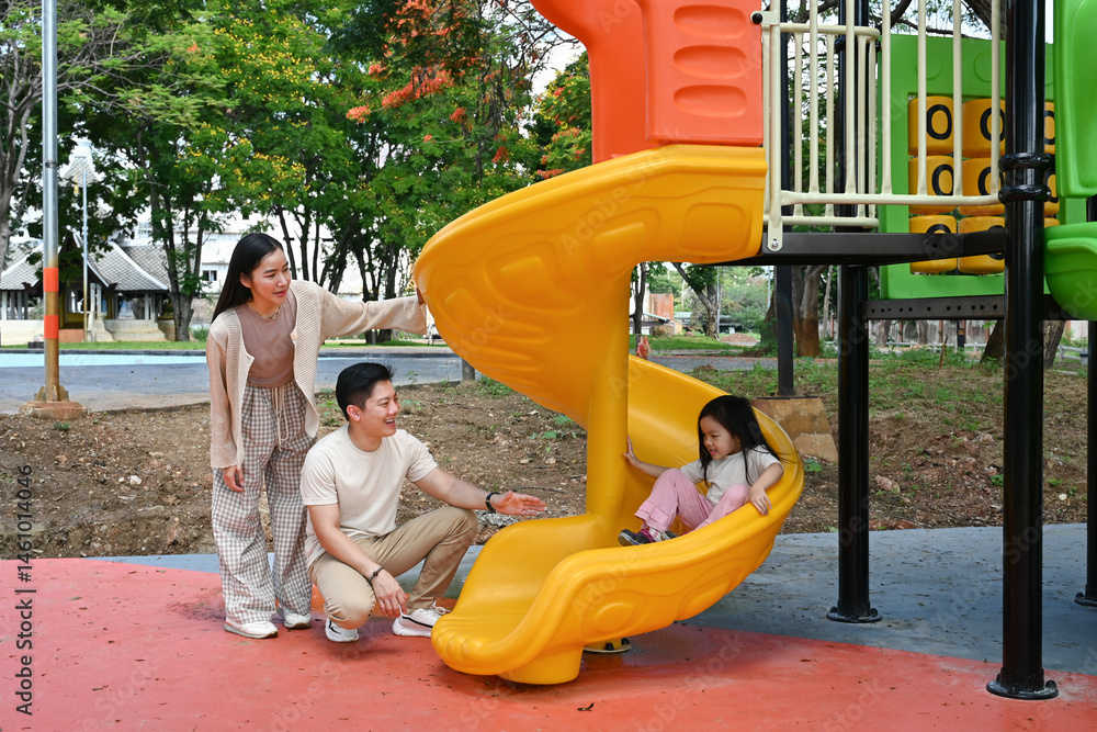 A family is standing in front of a slide. The child is sitting on the slide while the parents look on. Scene is happy and playful.