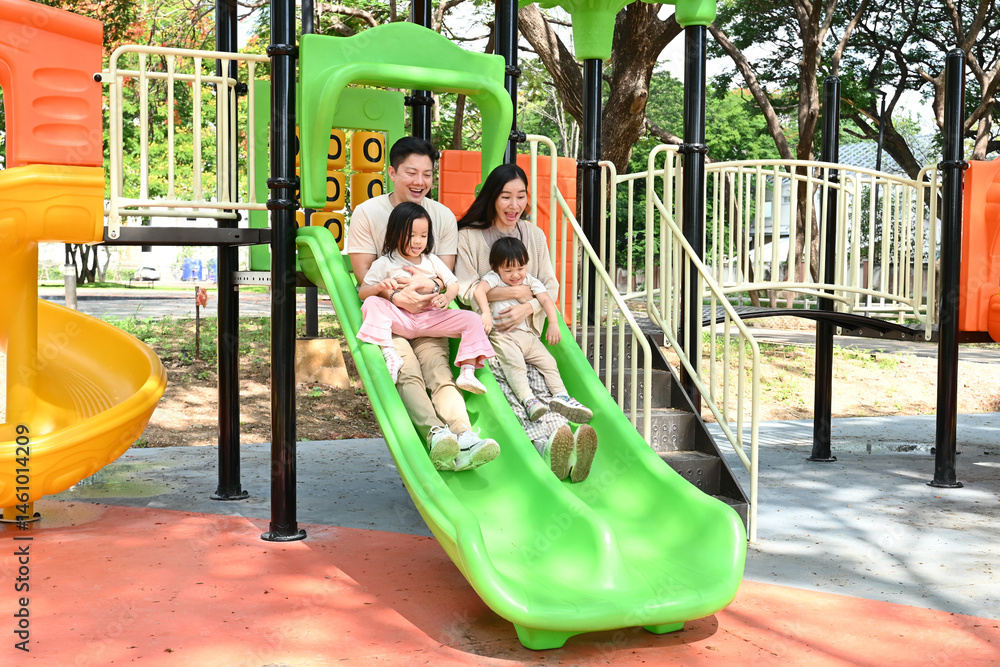 A family of four is enjoying a day at the park, riding a green slide together. Scene is happy and playful, as the family is spending quality time together and having fun.