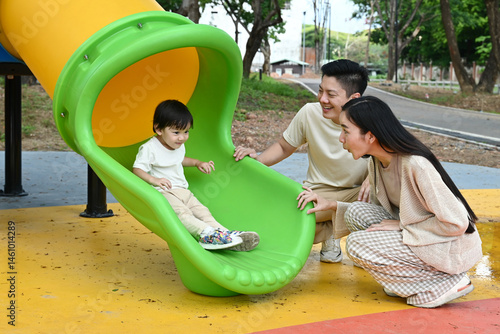 A family is standing in front of a slide. The child is sitting on the slide while the parents look on. Scene is happy and playful.