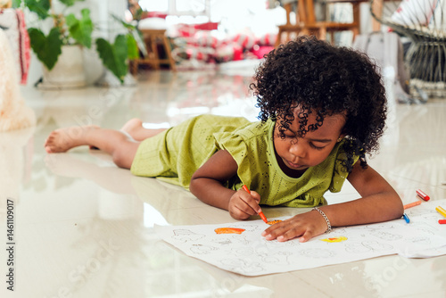 Photography Cute Black girl in green dress lying on floor, coloring with crayons on large paper