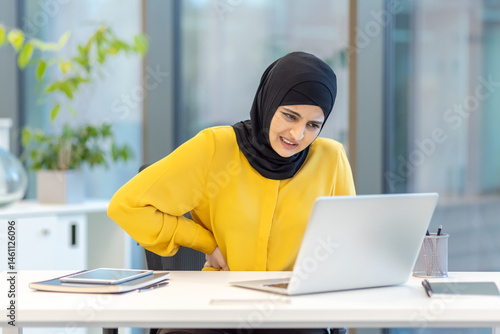 A woman wearing a hijab experiences back pain while working on her laptop at her desk in a modern office setting.