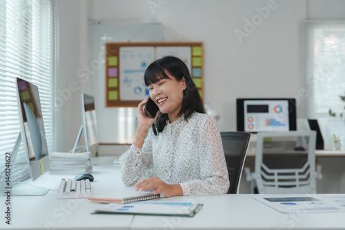 Cheerful young asian office employee sitting at a desk, engaging in a conversation on a smartphone while reviewing notes, surrounded by a computer and various documents