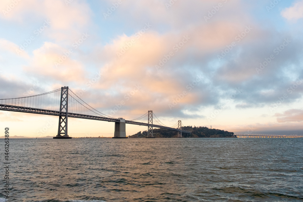 Fototapeta premium Bay Bridge stretching across the horizon beneath a soft sunset sky viewed from the San Francisco ferry on May 11, 2019