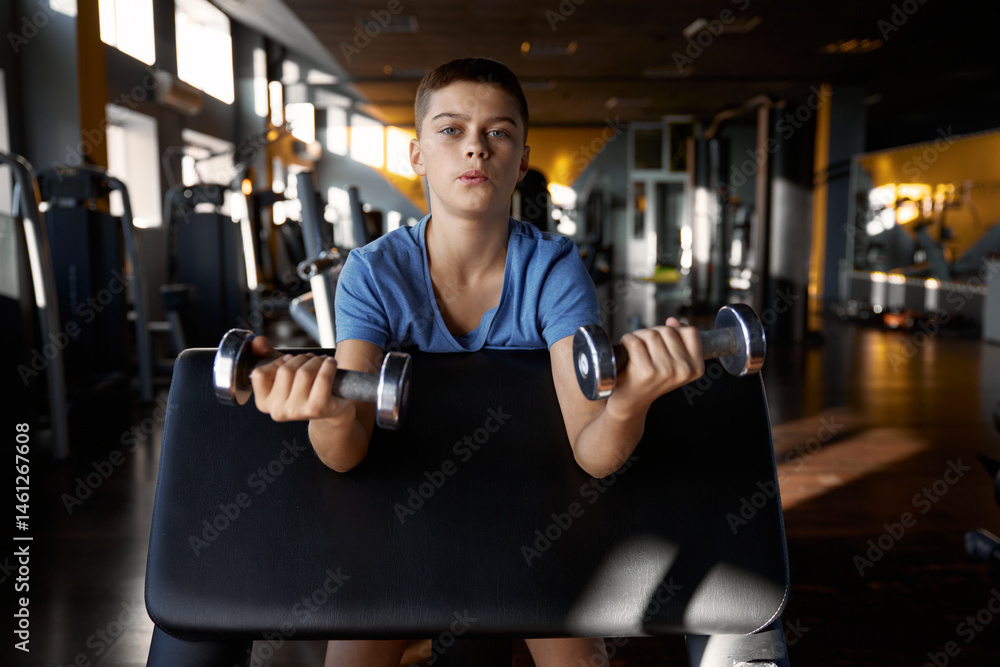 Teenage boy pumping biceps enjoying training with dumbbells at gym