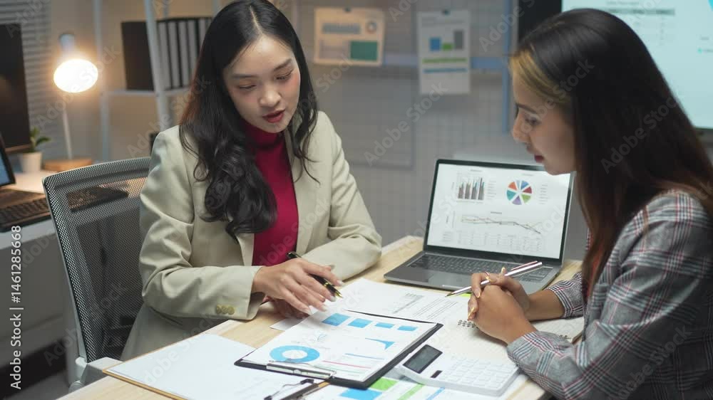 Two women are sitting at a desk with a white board and a laptop. One of them is writing on the white board Two women are sitting at a desk with a white board and a laptop. One of them is writing on the white board
