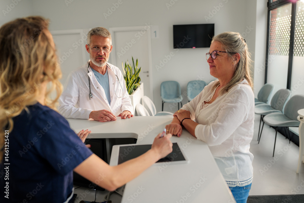 Obraz premium Mature female patient discussing treatment with a doctor while a nurse takes notes on a digital tablet at a modern hospital reception, highlighting collaborative healthcare