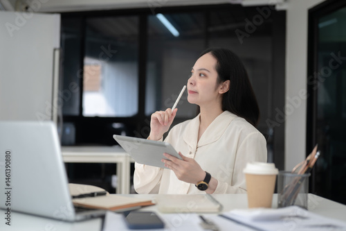 Businesswoman or office worker thinking while holding stylus and tablet sitting at table in office.