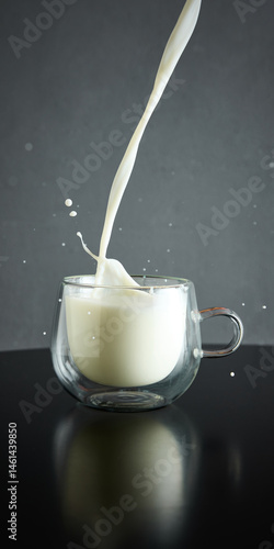 Milk pouring into clear glass mug, creating splash. Dark surface against grey background. Concept of drink, organic food, aesthetics, advertisement