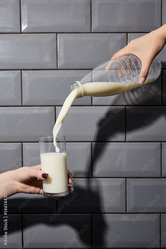 Hands hold plastic bottle and glass as milk pours with strong shadow on tiled wall. Concept of drink, organic food, aesthetics, advertisement