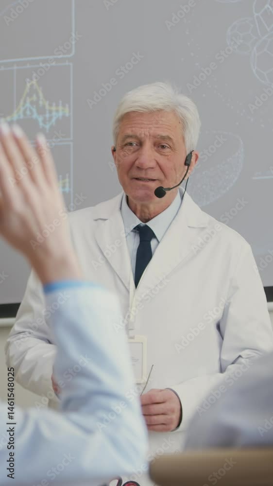 Vertical shot of senior male medical scholar equipped with earset microphone conducting lecture in auditorium then answering colleagues question during professional conference Vertical shot of senior male medical scholar equipped with earset microphone conducting lecture in auditorium then answering colleagues question during professional conference