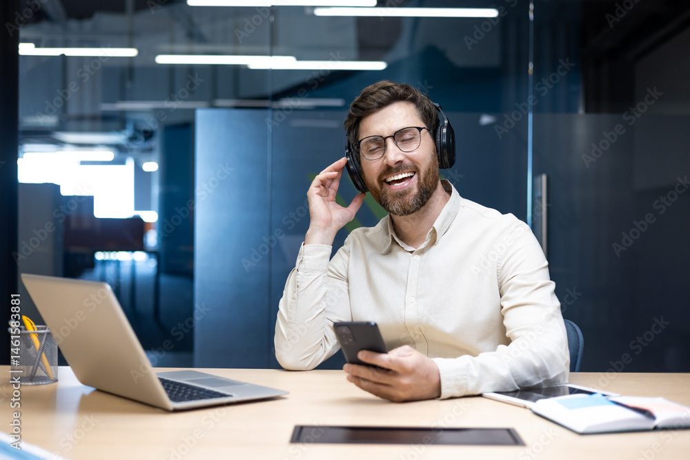 A smiling and relaxed young man is sitting at a desk in the office, wearing headphones, holding a phone in his hand and listening to music A smiling and relaxed young man is sitting at a desk in the office, wearing headphones, holding a phone in his hand and listening to music