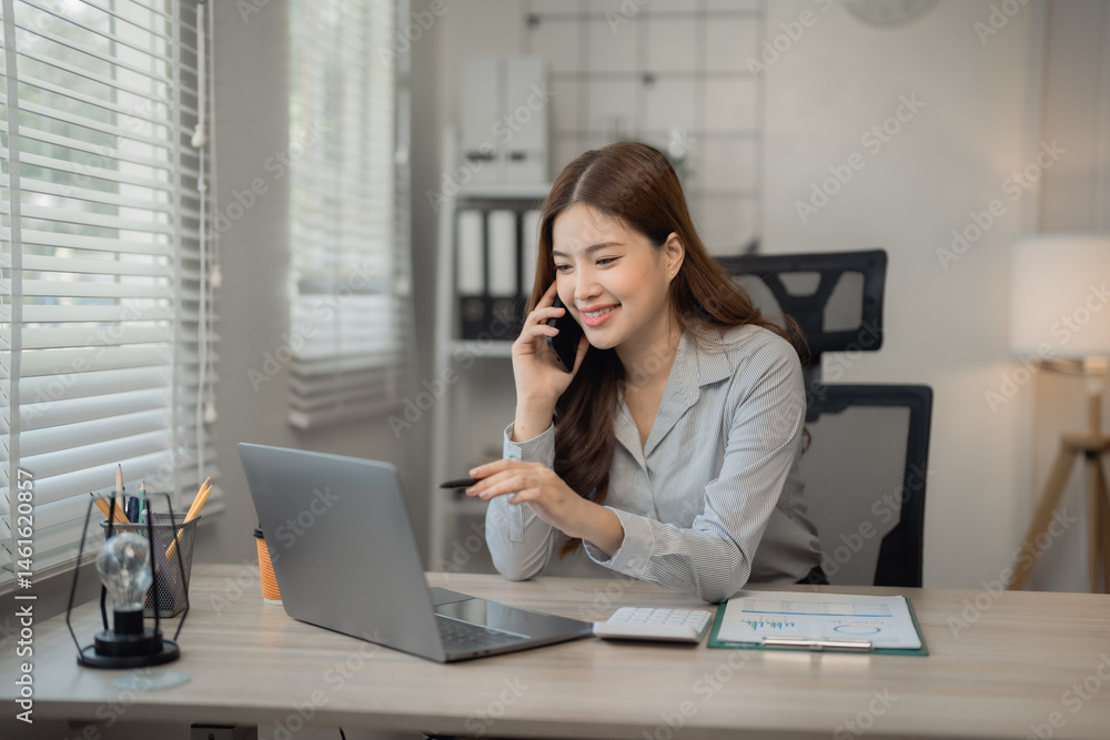 Young Asian businesswoman talking on mobile phone and pointing at laptop screen while working at modern office desk with documents, calculator and stationery Young Asian businesswoman talking on mobile phone and pointing at laptop screen while working at modern office desk with documents, calculator and stationery