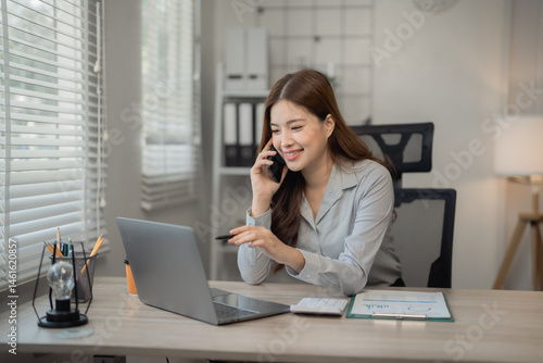 Young Asian businesswoman talking on mobile phone and pointing at laptop screen while working at modern office desk with documents, calculator and stationery