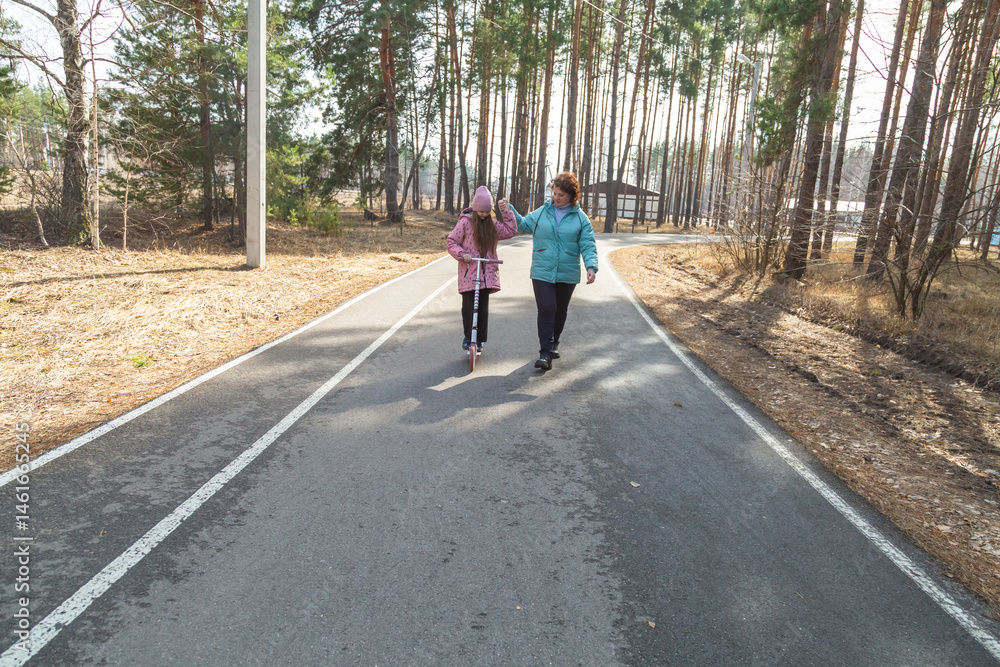 Fototapeta premium Little girl riding scooter while walking with her grandmother in the forest on a sunny day