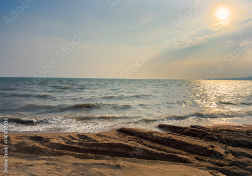 Coastal beach landscape with ocean waves and sunlight reflecting on water near sand covered in fresh vehicle tire tracks at golden sunset