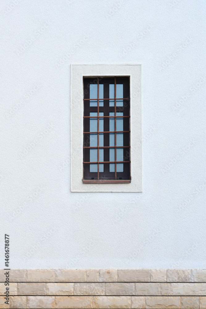 A small, dark window with multiple panes and metal bars is set in a textured white wall above a stone foundation